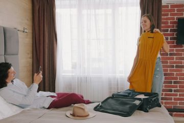 Žena drži žutu haljinu na vešalici dok muškarac sedi na krevetu i fotografiše je telefonom